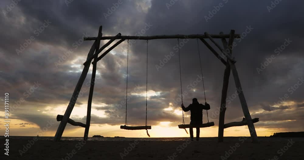 Travelling, vacation wanderlust concept. Silhouette of man tourist swinging on wooden swing on the sea beach at beautiful sunset, back view. Single man on a trip enjoys a vacation in nature. Travelling, vacation wanderlust concept. Silhouette of man tourist swinging on wooden swing on the sea beach at beautiful sunset, back view. Single man on a trip enjoys a vacation in nature.