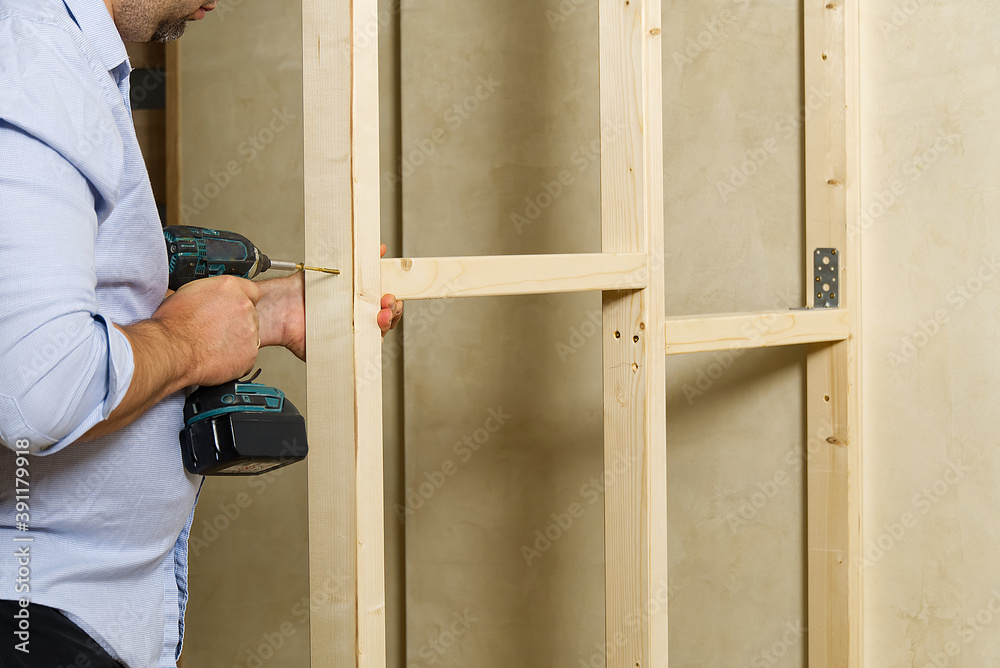 A man fastens two wooden beams with a self-tapping screw. The joiner wraps the screw with a screwdriver. Construction of a timber frame for plasterboard A man fastens two wooden beams with a self-tapping screw. The joiner wraps the screw with a screwdriver. Construction of a timber frame for plasterboard
