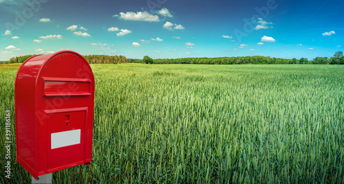 Fényképezés Big red modern postbox with white empty note space for address is standing outdoor in front of beautiful countryside landscape background with farm green wheat field and blue sky