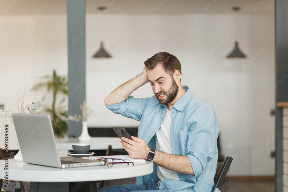 Puzzled man sit at table in coffee shop cafe working studying on laptop pc computer playing game on cell phone relaxing in restaurant during free time indoor. Freelance mobile office business concept. Puzzled man sit at table in coffee shop cafe working studying on laptop pc computer playing game on cell phone relaxing in restaurant during free time indoor. Freelance mobile office business concept.