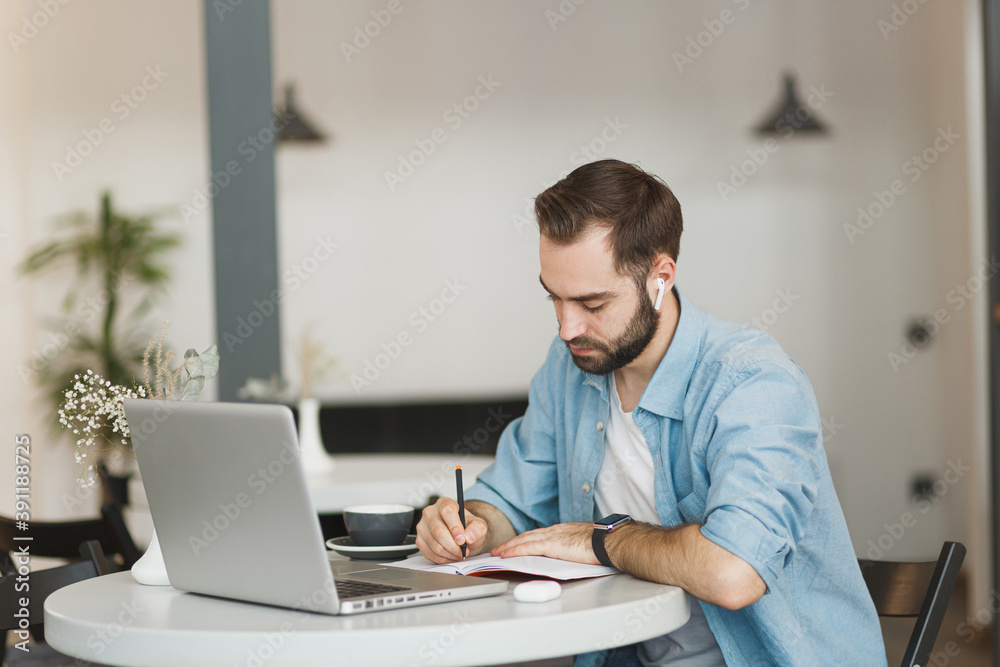 Handsome young man in air pods sitting alone at table in coffee shop cafe restaurant indoors working or studying on laptop pc computer writing in notebook. Freelance mobile office business concept. Handsome young man in air pods sitting alone at table in coffee shop cafe restaurant indoors working or studying on laptop pc computer writing in notebook. Freelance mobile office business concept.