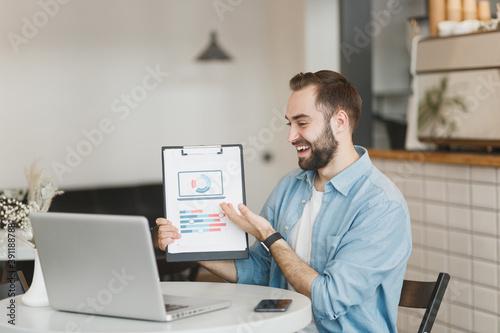 Cheerful young man sit at table in coffee shop cafe restaurant indoors working or studying on laptop pc computer pointing hand clipboard with papers document. Freelance mobile office business concept.