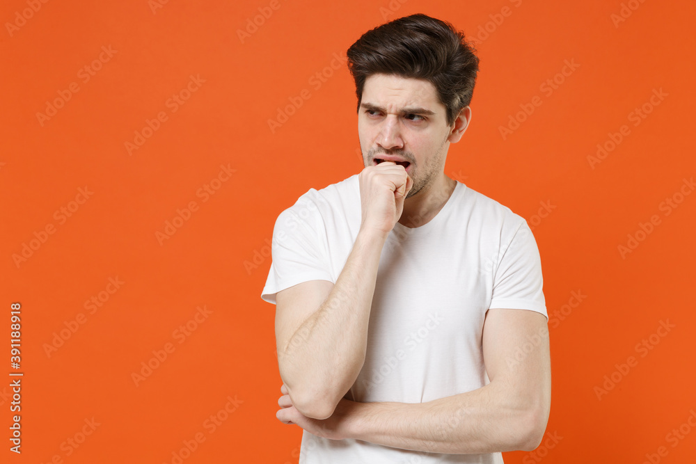 Preoccupied concerned worried nervous young man wearing basic casual empty blank white t-shirt standing gnawing nails looking aside isolated on bright orange colour wall background, studio portrait. Preoccupied concerned worried nervous young man wearing basic casual empty blank white t-shirt standing gnawing nails looking aside isolated on bright orange colour wall background, studio portrait.