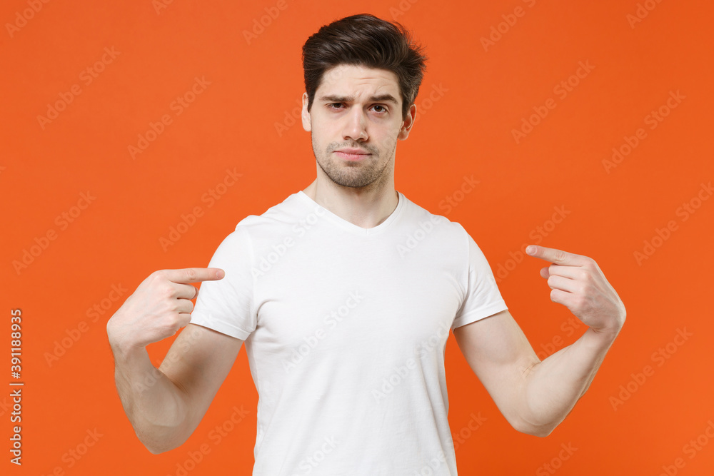 Perplexed unconfident young man 20s wearing basic casual white blank empty t-shirt standing pointing index fingers on himself looking camera isolated on bright orange wall background studio portrait. Perplexed unconfident young man 20s wearing basic casual white blank empty t-shirt standing pointing index fingers on himself looking camera isolated on bright orange wall background studio portrait.