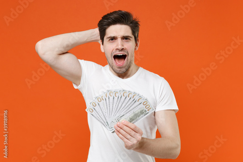 Fotografie Shocked amazed young man in basic casual empty white t-shirt hold fan of cash money in dollar banknotes put hand on head looking camera isolated on bright orange colour background studio portrait