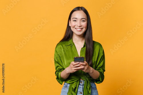Canvas Print Smiling cheerful young brunette asian woman wearing basic green shirt standing using mobile cell phone typing sms message looking camera isolated on bright yellow colour background, studio portrait