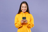 Smiling young brunette asian woman 20s wearing basic yellow hoodie standing holding in hands using mobile cell phone typing sms message isolated on pastel violet colour background, studio portrait.