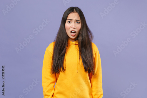 Shocked irritated worried young brunette asian woman 20s wearing casual basic yellow hoodie standing keeping mouth open looking aside isolated on pastel violet colour background studio portrait.