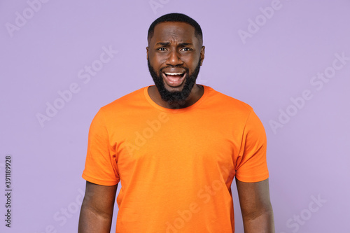 Shocked worried nervous young african american man 20s wearing basic casual orange t-shirt standing keeping mouth open looking camera isolated on pastel violet colour background, studio portrait.