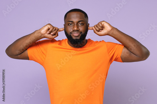 Dissatisfied displeased confused young african american man wearing basic casual empty orange t-shirt covering ears with fingers looking camera isolated on pastel violet background studio portrait.