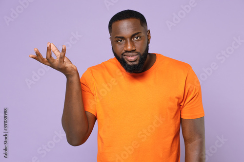 Confused smirked puzzled young african american man 20s wearing basic casual blank empty orange t-shirt spreading hands looking camera isolated on pastel violet colour background studio portrait.