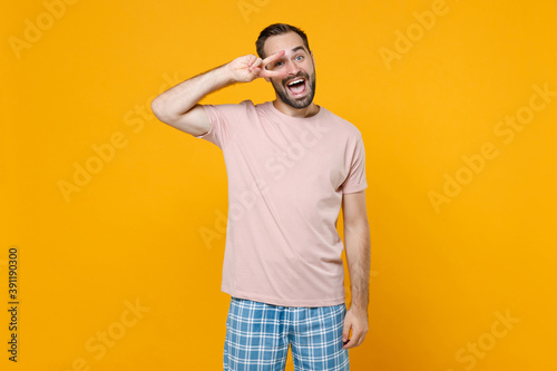 Excited young bearded man in pajamas home wear showing victory sign looking camera while resting at home isolated on bright yellow colour background studio portrait. Relax good mood lifestyle concept.