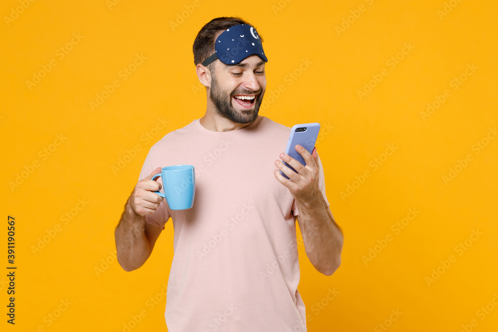 Cheerful young man in pajamas home wear sleep mask hold cup coffee tea drinking using mobile cell phone rest at home isolated on yellow background studio portrait. Relax good mood lifestyle concept. Cheerful young man in pajamas home wear sleep mask hold cup coffee tea drinking using mobile cell phone rest at home isolated on yellow background studio portrait. Relax good mood lifestyle concept.