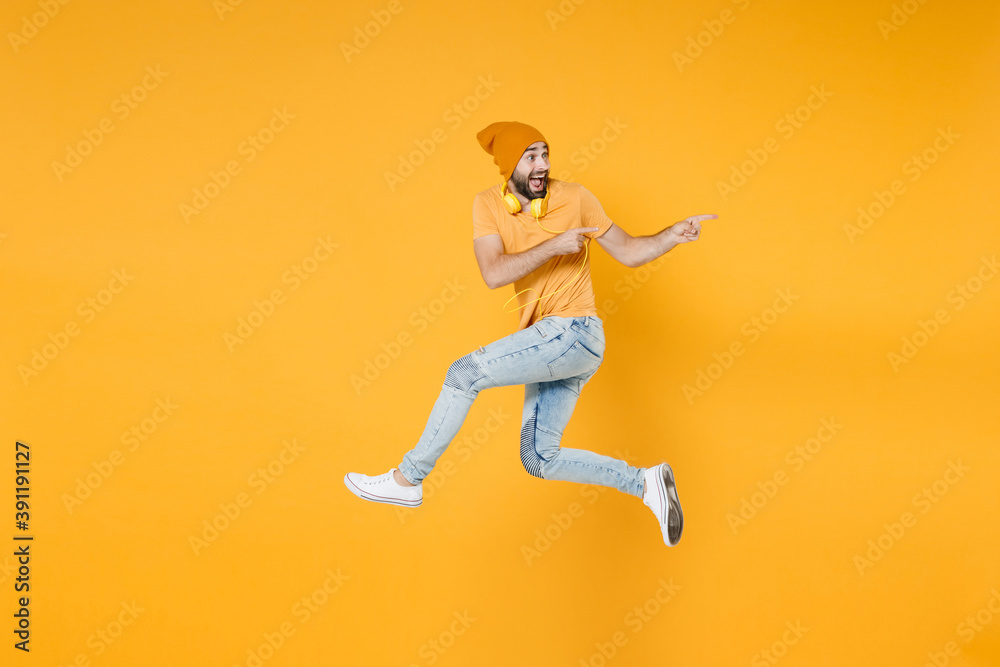 Full length side view of shocked young man wearing basic casual t-shirt headphones hat jumping pointing index fingers aside on mock up copy space isolated on bright yellow background, studio portrait.