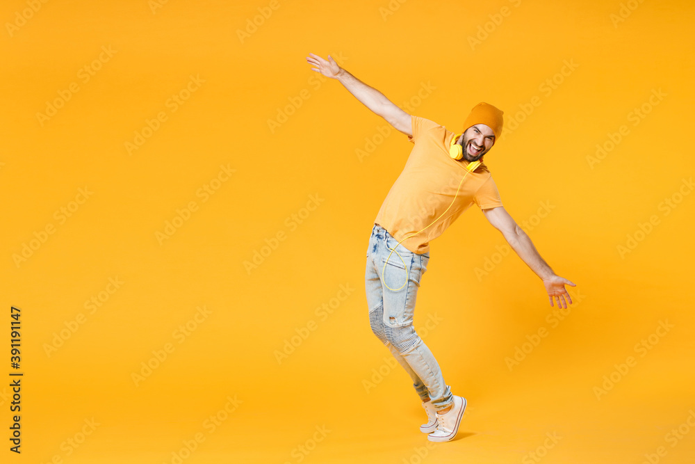 Full length side view of laughing funny young man in basic casual t-shirt headphones hat posing dancing standing on toes spreading hands looking camera isolated on yellow background studio portrait.