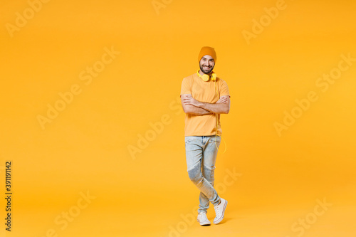 Full length of cheerful smiling young man 20s wearing basic casual t-shirt headphones hat standing holding hands crossed looking camera isolated on bright yellow colour background, studio portrait.