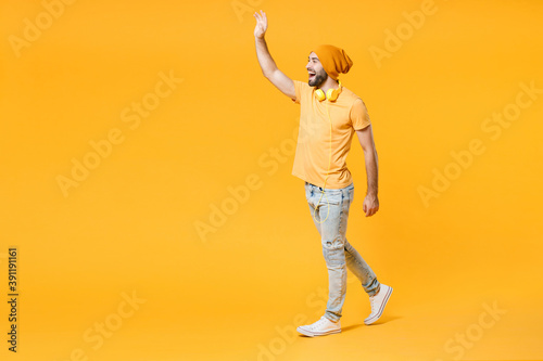 Full length side view of cheerful young man wearing basic casual t-shirt headphones hat waving and greeting with hand as notices someone looking aside isolated on yellow background, studio portrait.