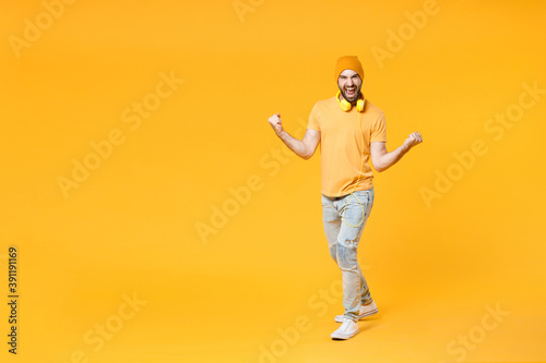 Full length of overjoyed screaming young man in basic casual t-shirt headphones hat standing clenching fists doing winner gesture looking camera isolated on bright yellow background, studio portrait.