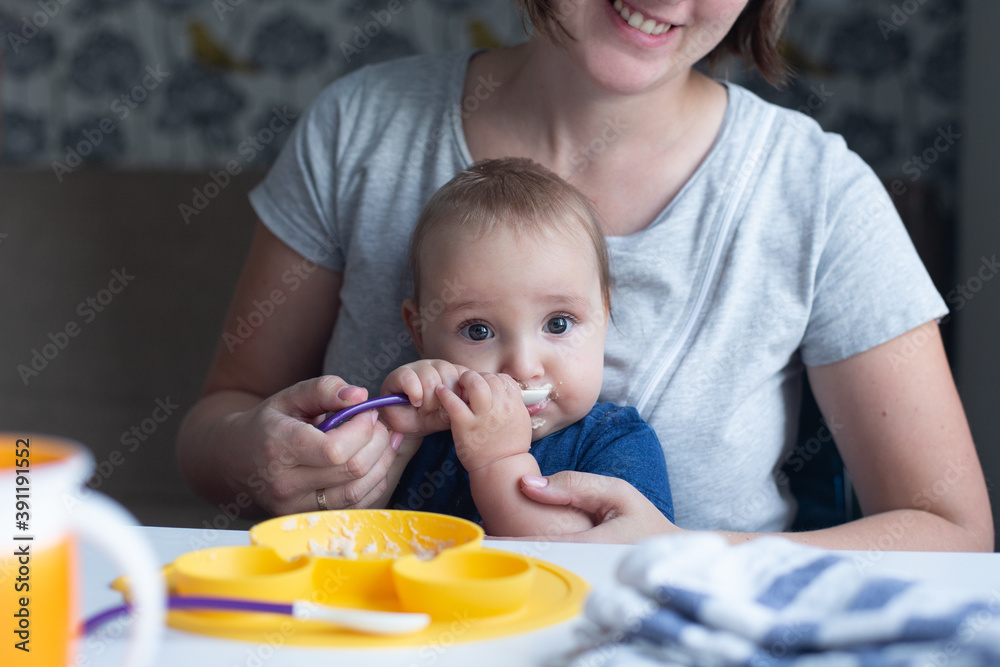 Cute infant trying to eat baby food using spoon and his cheerful mother assisting him. Cute infant trying to eat baby food using spoon and his cheerful mother assisting him.