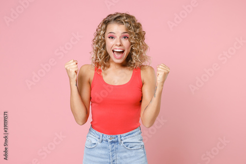 Happy joyful excited shocked young blonde woman wearing basic casual tank top standing clenching fists doing winner gesture looking camera isolated on pastel pink colour background, studio portrait.