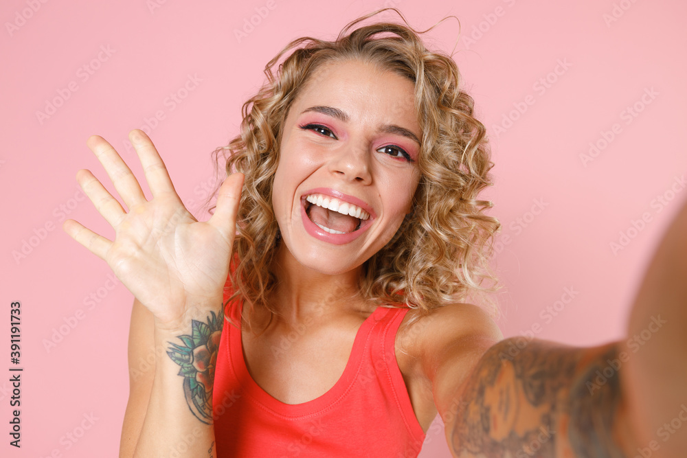 Close up of excited young blonde woman in basic casual tank top standing doing selfie shot on mobile phone waving greeting with hand isolated on pastel pink colour wall background, studio portrait. Close up of excited young blonde woman in basic casual tank top standing doing selfie shot on mobile phone waving greeting with hand isolated on pastel pink colour wall background, studio portrait.