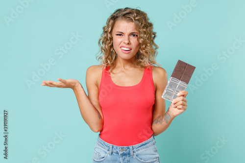 Confused puzzled worried young blonde woman wearing pink basic casual tank top standing hold chocolate bar spreading hands looking camera isolated on blue turquoise colour background studio portrait.