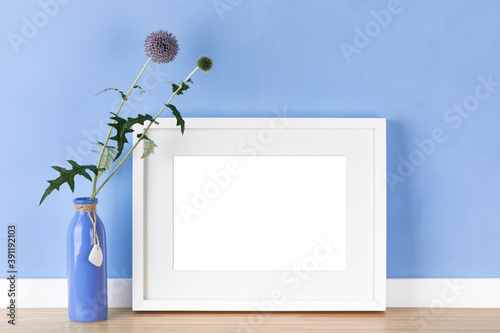 Elegant horizontal white picture frame with matte poster artwork mockup, template for online shop with globe thistle flower in front of pastel blue wall. Blank image area isolated with clipping path.