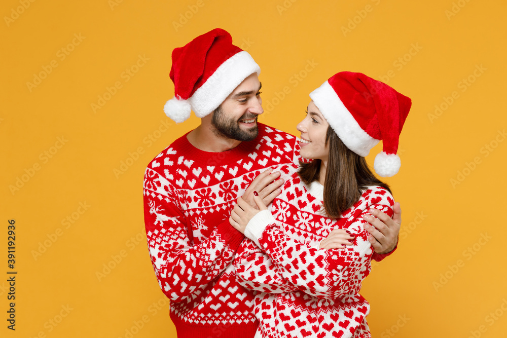 Smiling young Santa couple friends man woman 20s wearing red sweater Christmas hat looking at each other hugging isolated on yellow background studio. Happy New Year celebration merry holiday concept. Smiling young Santa couple friends man woman 20s wearing red sweater Christmas hat looking at each other hugging isolated on yellow background studio. Happy New Year celebration merry holiday concept.