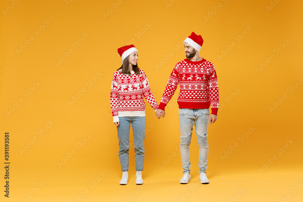 Obraz premium Full length of smiling young Santa couple friends man woman in sweater, Christmas hat hold hands looking at each other isolated on yellow background. Happy New Year celebration merry holiday concept.