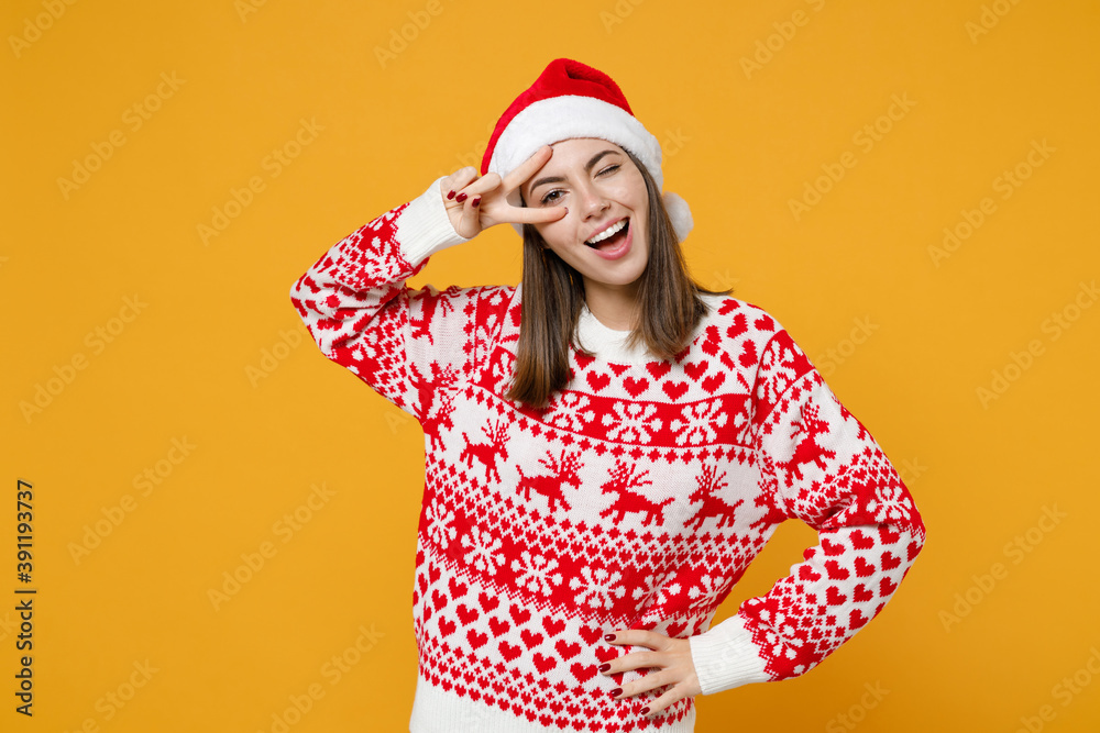 Blinking young brunette Santa woman in red sweater Christmas hat showing victory sign looking camera isolated on yellow background, studio portrait. Happy New Year celebration merry holiday concept. Blinking young brunette Santa woman in red sweater Christmas hat showing victory sign looking camera isolated on yellow background, studio portrait. Happy New Year celebration merry holiday concept.