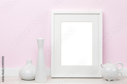 Elegant poster artwork mockup template for online shop with white vertical picture frame with matte, vases and tea kettle in front of pink wall. Blank image area isolated with clipping path.