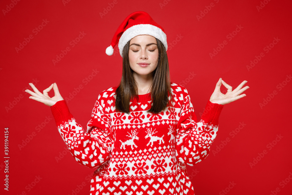 Relaxed young Santa woman in sweater Christmas hat hold hands in yoga gesture, relaxing meditating, trying to calm down isolated on red background. Happy New Year celebration merry holiday concept. Relaxed young Santa woman in sweater Christmas hat hold hands in yoga gesture, relaxing meditating, trying to calm down isolated on red background. Happy New Year celebration merry holiday concept.