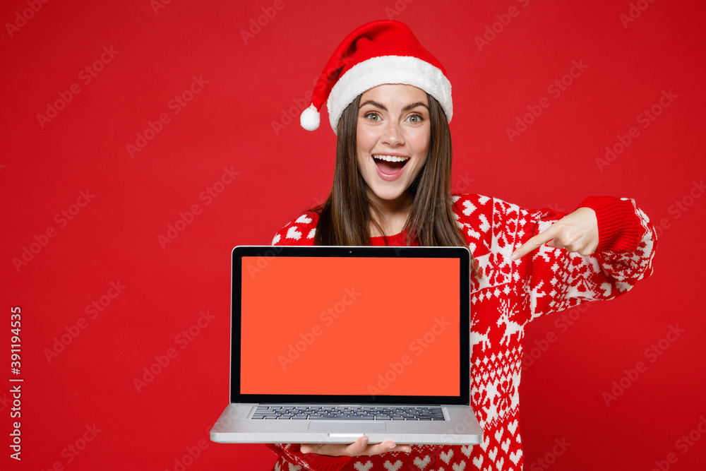 Excited young Santa woman in sweater Christmas hat point index finger on laptop pc computer with blank empty screen isolated on red background studio. Happy New Year celebration merry holiday concept. Excited young Santa woman in sweater Christmas hat point index finger on laptop pc computer with blank empty screen isolated on red background studio. Happy New Year celebration merry holiday concept.