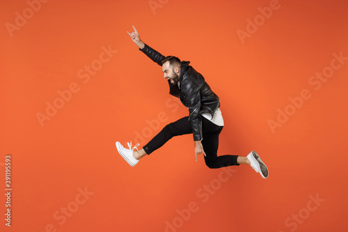 Full length side view of crazy screaming young bearded man in white t-shirt black leather jacket jumping depicting heavy metal rock sign isolated on bright orange colour background studio portrait.