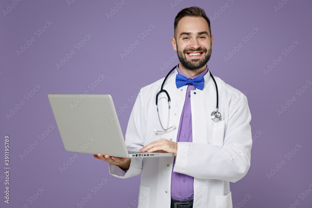 Smiling young bearded doctor man in white medical gown stethoscope working on laptop pc computer isolated on violet colour background studio portrait. Healthcare personnel health medicine concept. Smiling young bearded doctor man in white medical gown stethoscope working on laptop pc computer isolated on violet colour background studio portrait. Healthcare personnel health medicine concept.