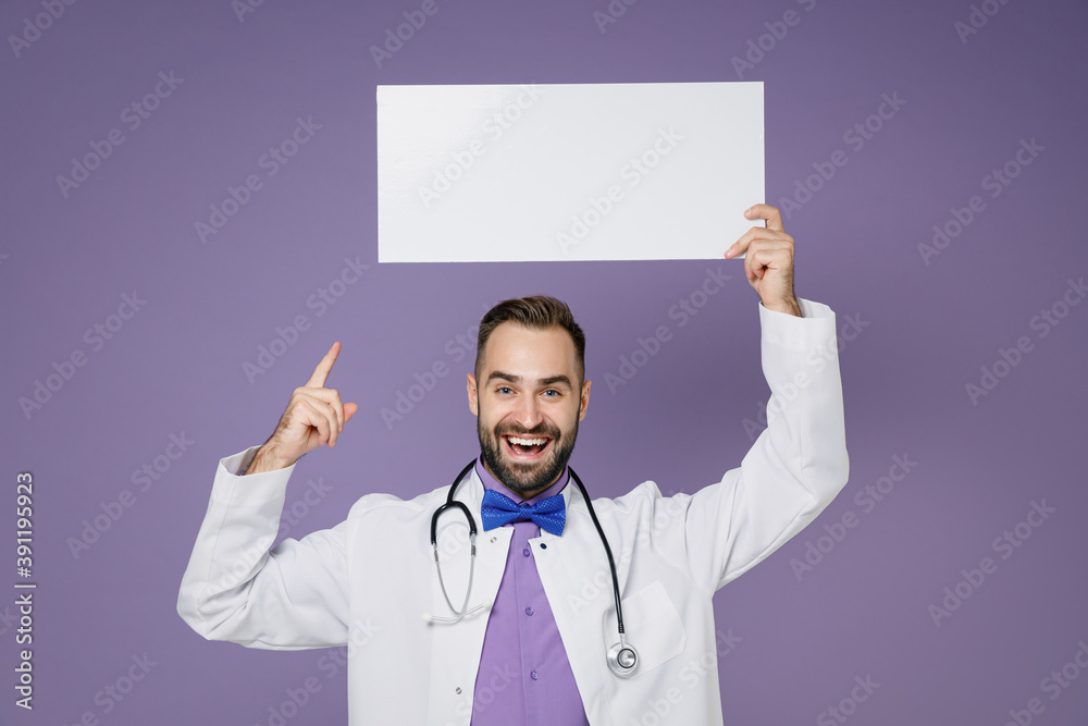Funny young bearded doctor man in medical gown stethoscope pointing index finger on blank sign board with place for text isolated on violet background. Healthcare personnel health medicine concept. Funny young bearded doctor man in medical gown stethoscope pointing index finger on blank sign board with place for text isolated on violet background. Healthcare personnel health medicine concept.