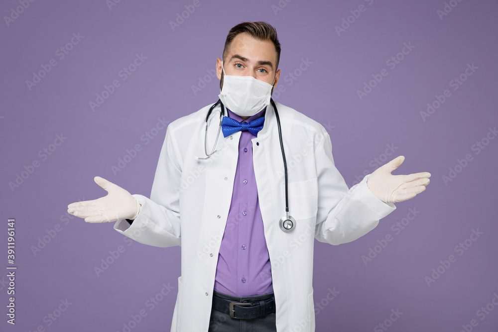 Confused young doctor man in medical gown stethoscope face mask to safe from coronavirus virus covid-19 spreading hands isolated on violet background. Healthcare personnel health medicine concept.