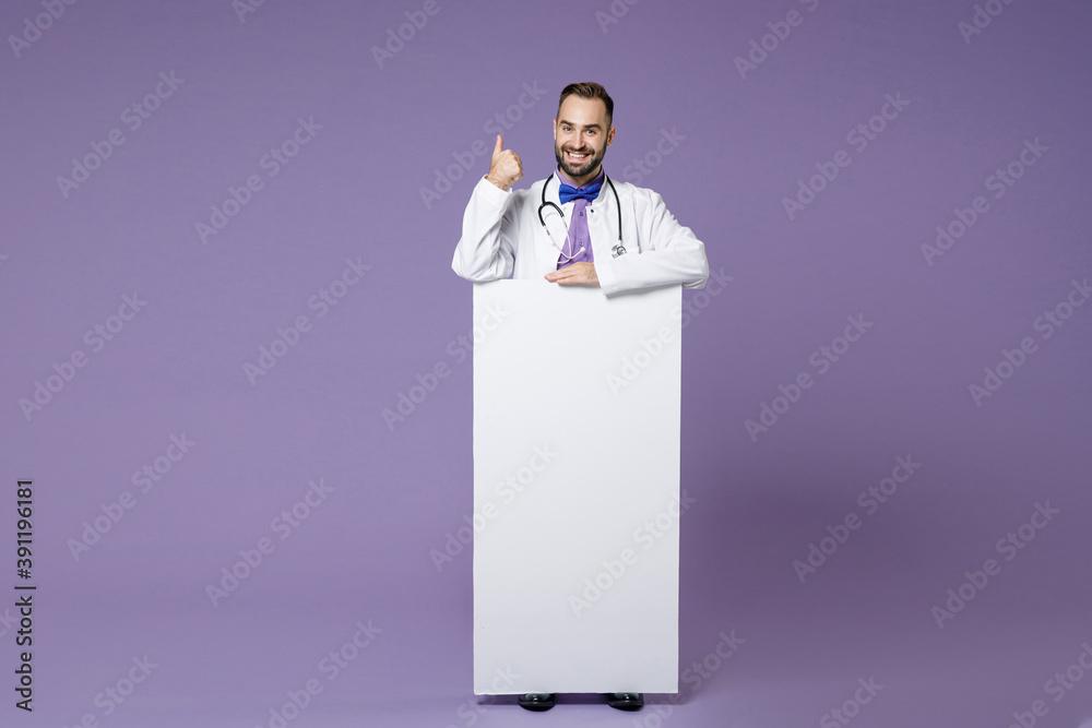 Full length of smiling young bearded doctor man in white medical gown hold big white empty blank billboard showing thumb up isolated on violet background. Healthcare personnel health medicine concept. Full length of smiling young bearded doctor man in white medical gown hold big white empty blank billboard showing thumb up isolated on violet background. Healthcare personnel health medicine concept.