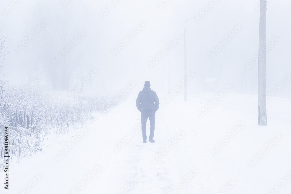 One young adult man in black clothes walking on sidewalk in heavy mist in white winter day. Foggy air. Poor visibility. Back view. One young adult man in black clothes walking on sidewalk in heavy mist in white winter day. Foggy air. Poor visibility. Back view.