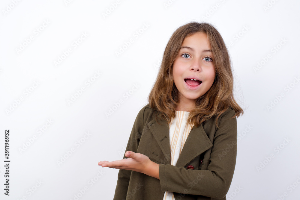 Beautiful little girl standing against white background, says: wow how exciting it is, has amazed expression, shows something on blank space with palm. Advertisement concept. Beautiful little girl standing against white background, says: wow how exciting it is, has amazed expression, shows something on blank space with palm. Advertisement concept.