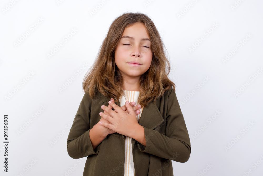 Beautiful little girl standing against white backgr closes eyes and keeps hands on chest near heart, expresses sincere emotions, being kind hearted and honest. Body language and real feelings concept. Beautiful little girl standing against white backgr closes eyes and keeps hands on chest near heart, expresses sincere emotions, being kind hearted and honest. Body language and real feelings concept.