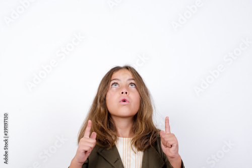 Photos Beautiful little girl standing against white background,  being amazed and surprised looking and pointing up with fingers showing something strange