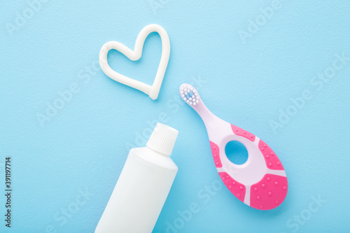 White heart shape, tube of toothpaste, pink toothbrush on light blue table background. Pastel color. Babies teeth hygiene. Closeup. Top down view.
