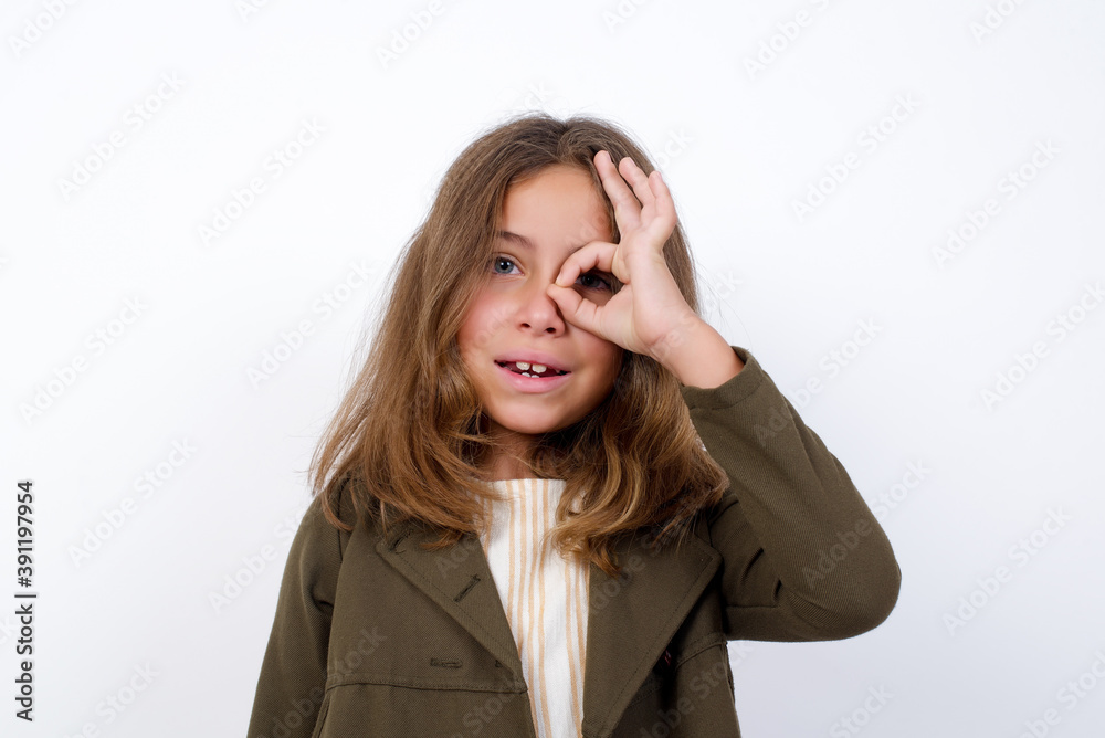 Beautiful little girl standing against white background, with happy face smiling doing ok sign with hand on eye looking through finger. Beautiful little girl standing against white background, with happy face smiling doing ok sign with hand on eye looking through finger.