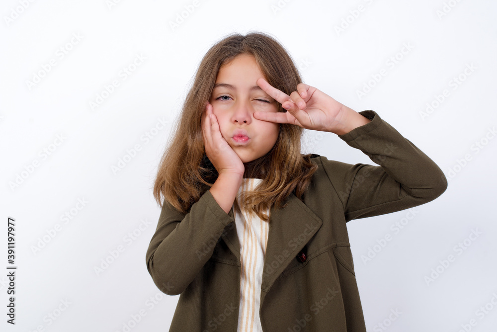 Beautiful little girl standing against white background, making v-sign near eyes. Leisure lifestyle people person celebrate flirt coquettish concept. Beautiful little girl standing against white background, making v-sign near eyes. Leisure lifestyle people person celebrate flirt coquettish concept.