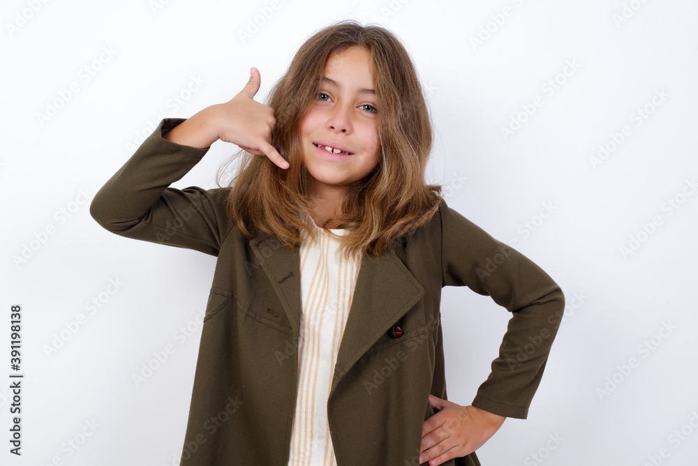 Beautiful little girl standing against white background, imitates telephone conversation, makes phone call gesture with hands, has confident expression. Call me! Beautiful little girl standing against white background, imitates telephone conversation, makes phone call gesture with hands, has confident expression. Call me!