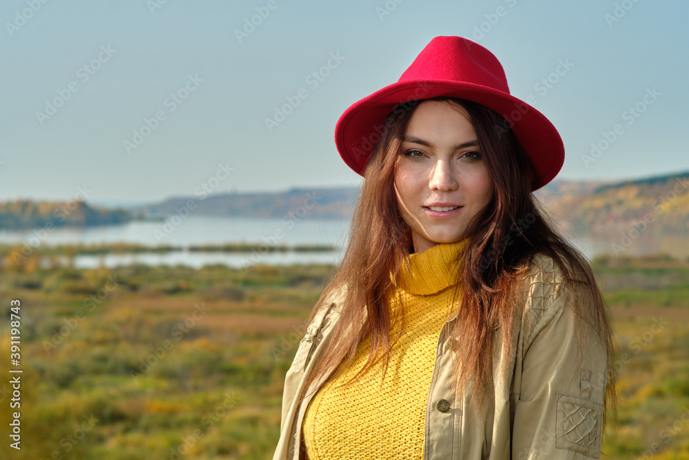 a girl in a yellow sweater raincoat and a red hat stands on the background of the river in the autumn landscape alone