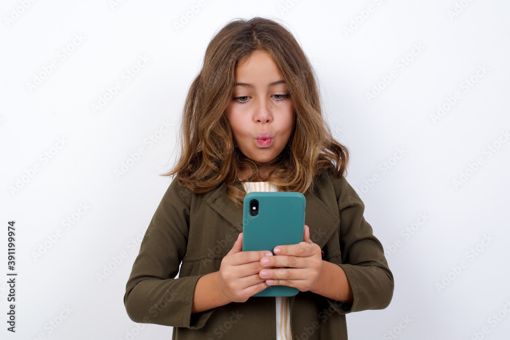 Beautiful little girl standing against white background, looks with bugged eyes, holds modern smart phone, receives unexpected message from friend, reads reminder. Beautiful little girl standing against white background, looks with bugged eyes, holds modern smart phone, receives unexpected message from friend, reads reminder.