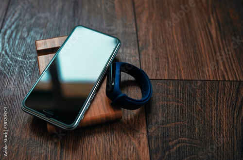 A cell phone (buttonless) and a smart bracelet lie on a wooden table stand. The light from the window hits the screen at an angle.