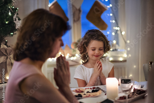 Senior woman with small granddaughter indoors at Christmas, praying at the table.